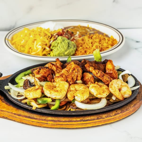 Sizzling fajitas with shrimp, chicken, onions, and peppers, alongside a plate of rice, beans, and guacamole.