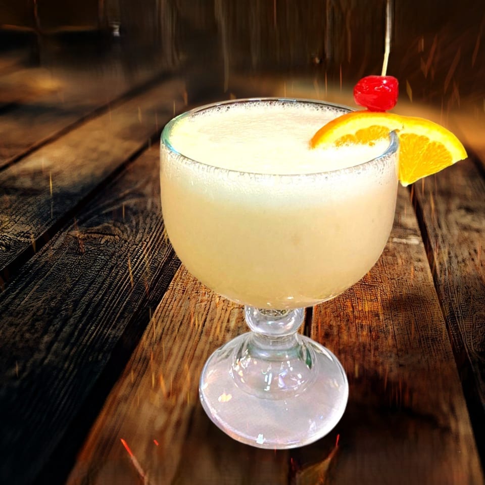 A frozen margarita in a goblet, garnished with an orange slice and a cherry, on a wooden table.
