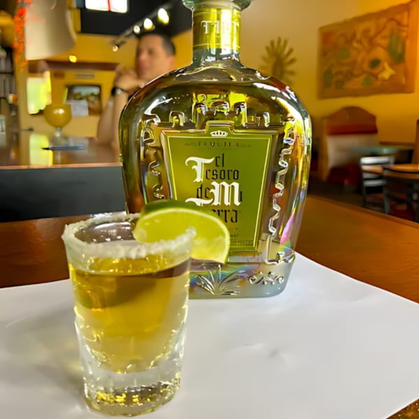 A shot of tequila with a lime wedge and a large bottle of El Tesoro de Tierra tequila.
